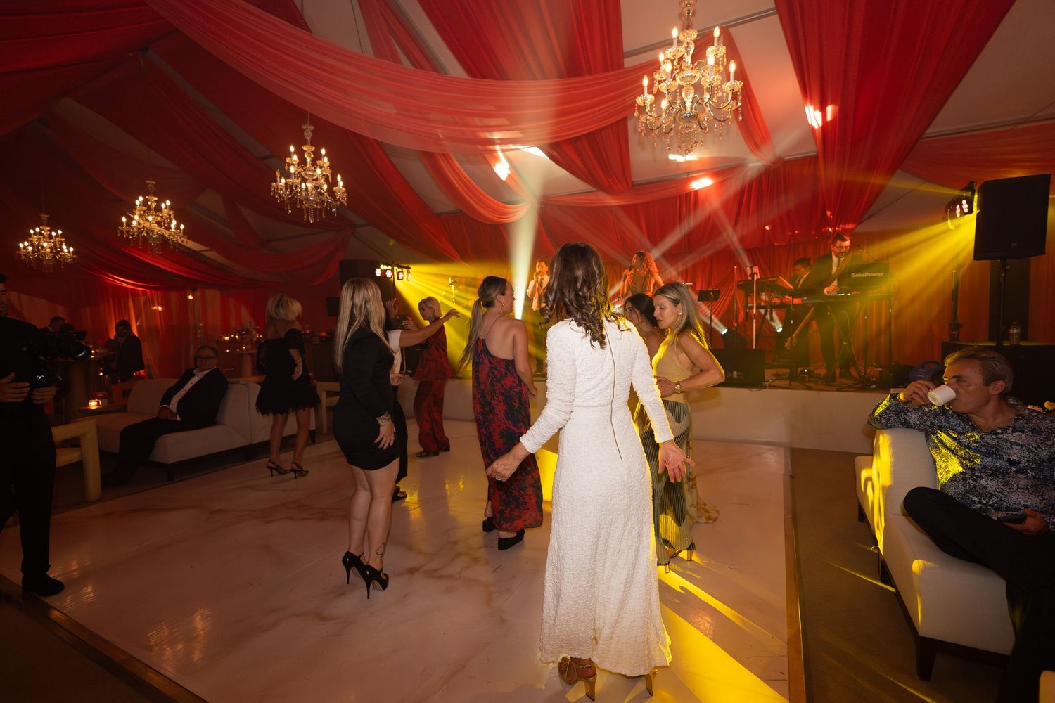 People dancing at a wedding reception with red draping decor and chandeliers overhead.