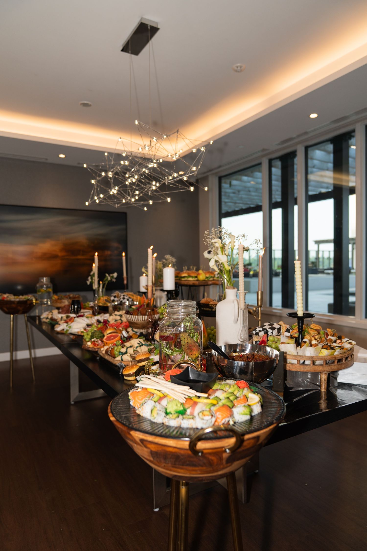 Elegant catering display with gourmet hors d'oeuvres under modern crystal chandelier in upscale event space with city views.