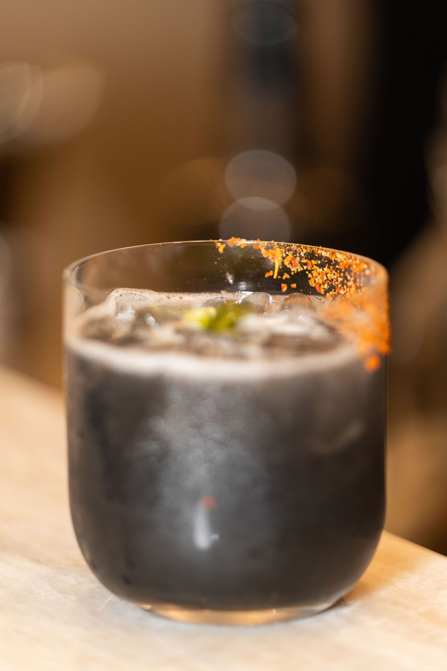 A dark cocktail served in a rocks glass with a gold-rimmed garnish and ice cubes in moody lighting.