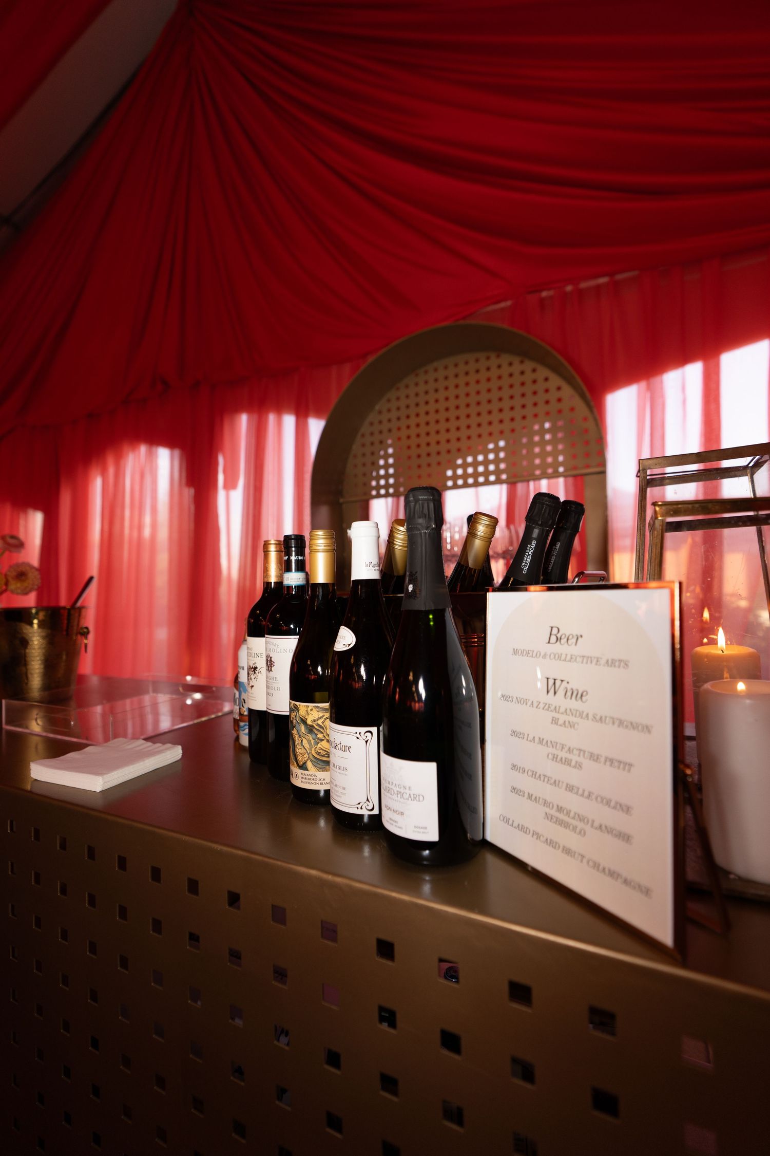 Wine bottles lined up on a wooden shelf against red draped fabric in a dimly lit display.