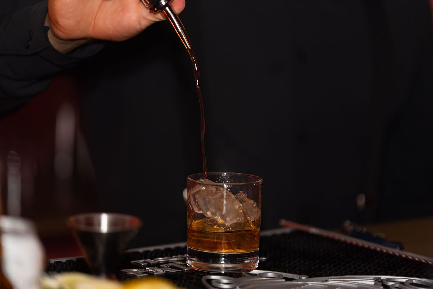 A hand pours dark liquid into a rocks glass with ice cubes on a black surface.