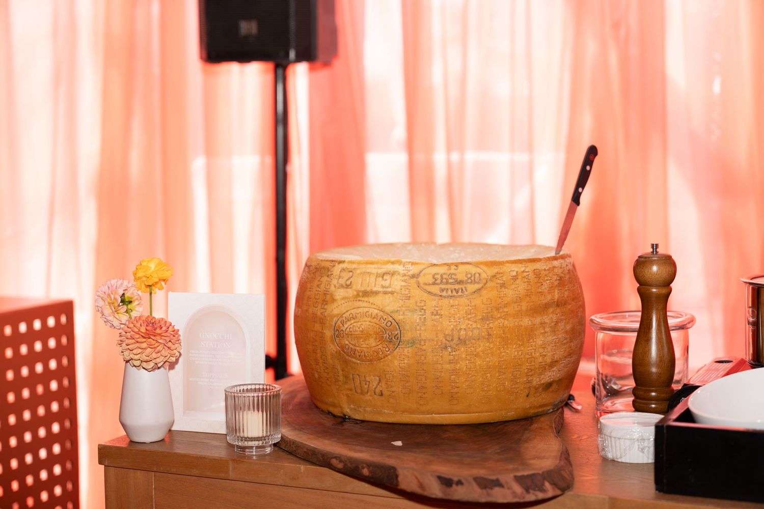 A large wheel of aged Parmesan cheese sits on a wooden board with a pepper mill and small vase of flowers nearby.