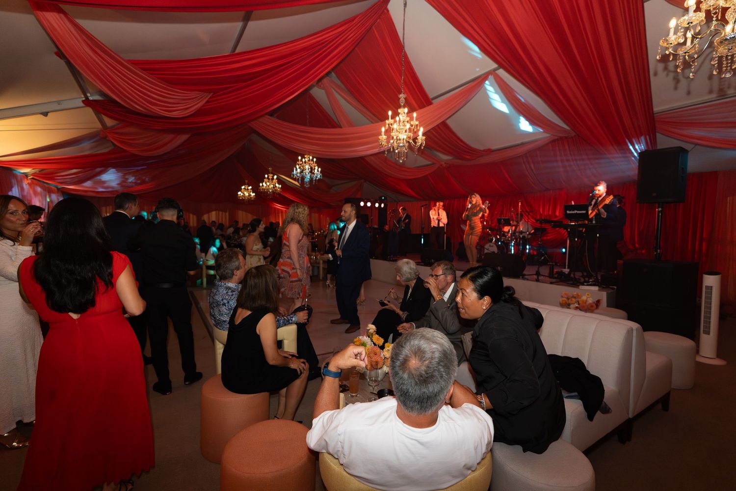 Red draped fabric decorates the ceiling of an elegant reception venue with guests mingling and dancing.