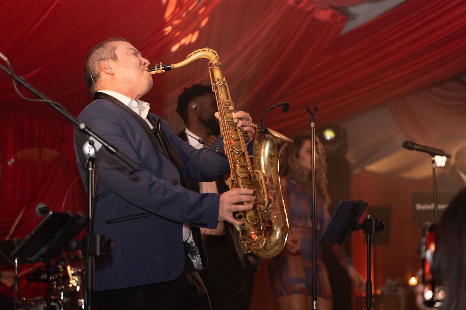 A musician plays saxophone on stage with dramatic red lighting and decor in the background.