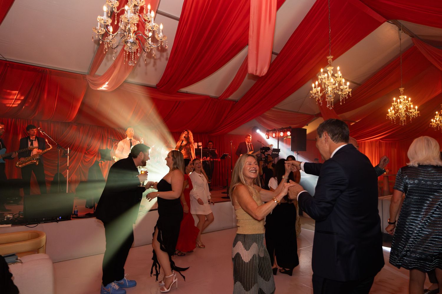 A wedding reception venue decorated with red uplighting and crystal chandeliers as guests dance on the dance floor.