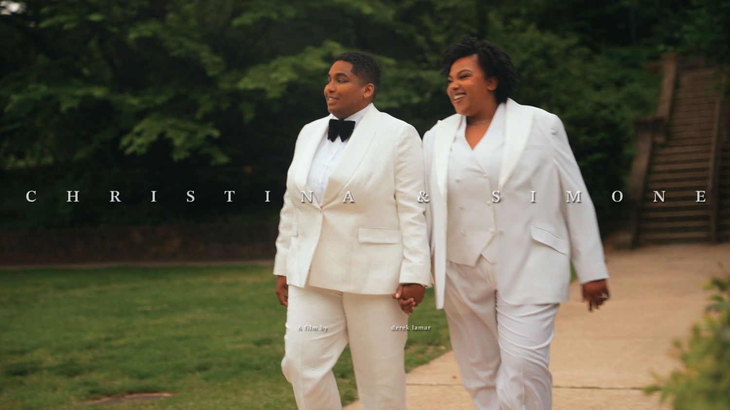 Same-sex couple Christina and Simone walking hand in hand in matching white wedding suits outdoors.