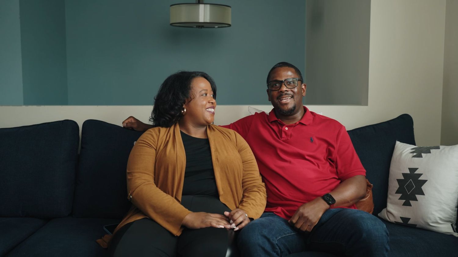 A smiling couple sitting together on a navy blue couch in a modern living room with teal walls.