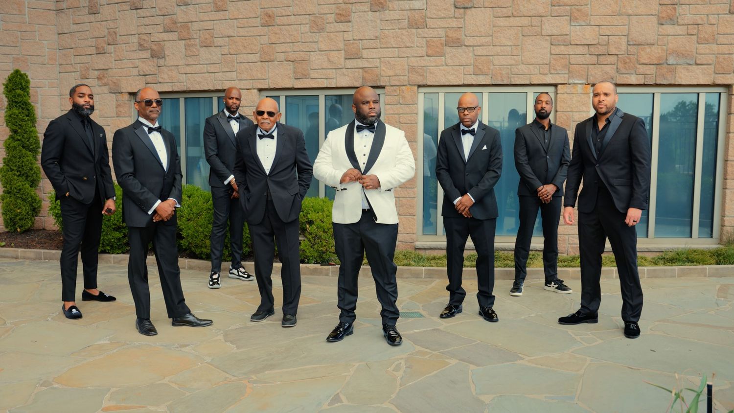 Groom in white tuxedo posed with seven groomsmen in black suits outside a modern building.