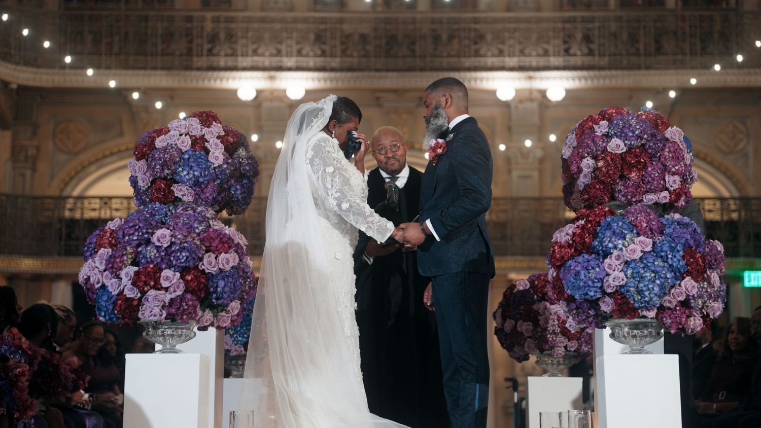 Couple exchanges vows at indoor wedding ceremony surrounded by lush floral arrangements.