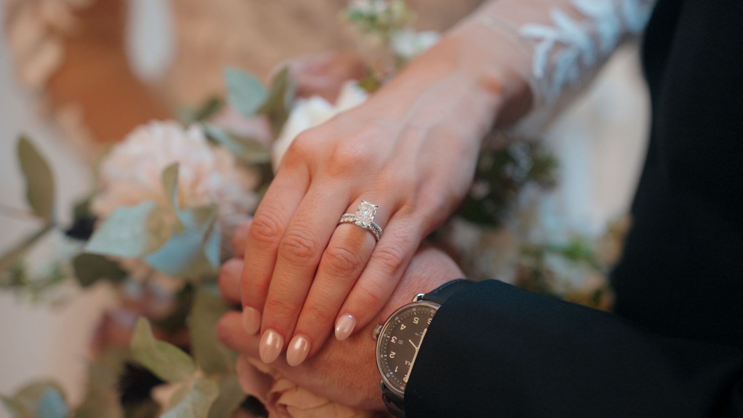 Close-up of bride and groom hands showing diamond engagement ring and wedding bands with floral backdrop.