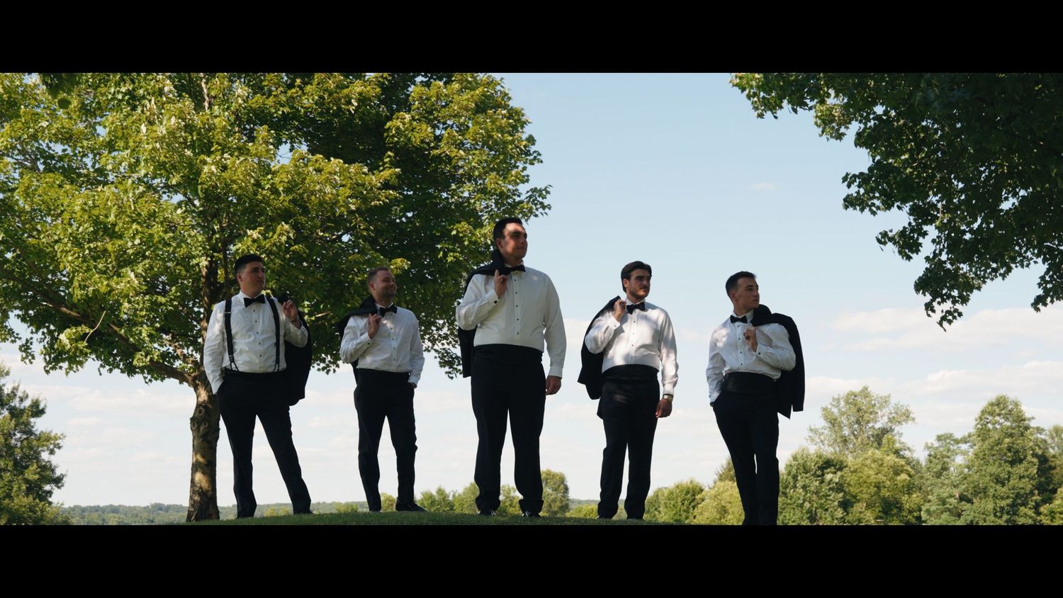 Five groomsmen in black pants and white shirts with suspenders standing outdoors on a sunny day.