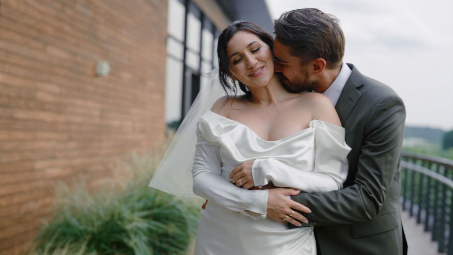 A groom in a gray suit embraces his bride in a white off-shoulder gown outdoors on their wedding day.