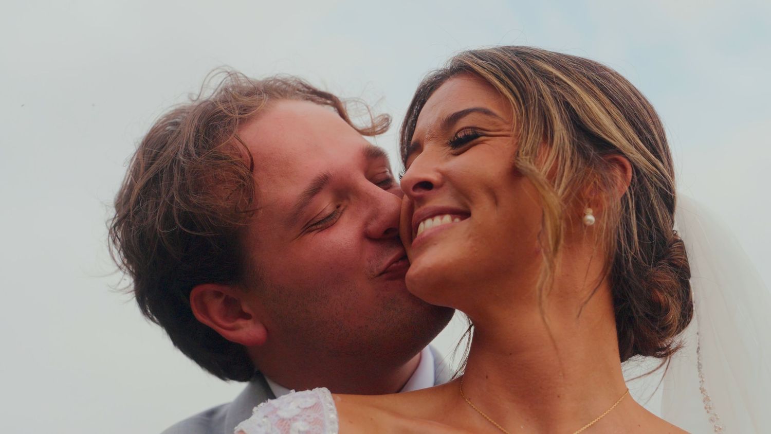 A romantic couple sharing a kiss outdoors, with the man kissing the woman's cheek as she smiles warmly.