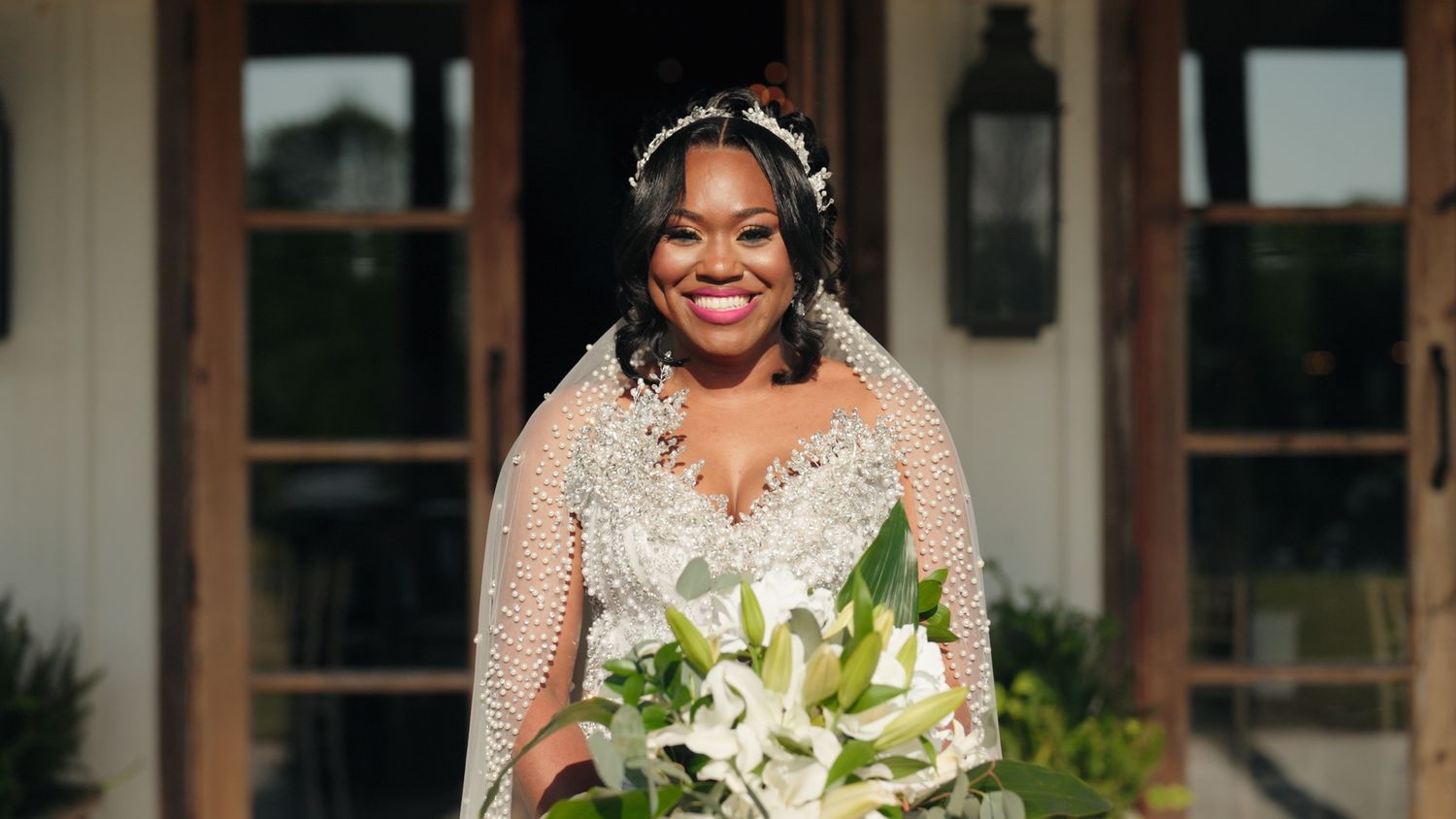 Radiant bride smiling in a beaded wedding gown and floral crown holding white lily bouquet outdoors.