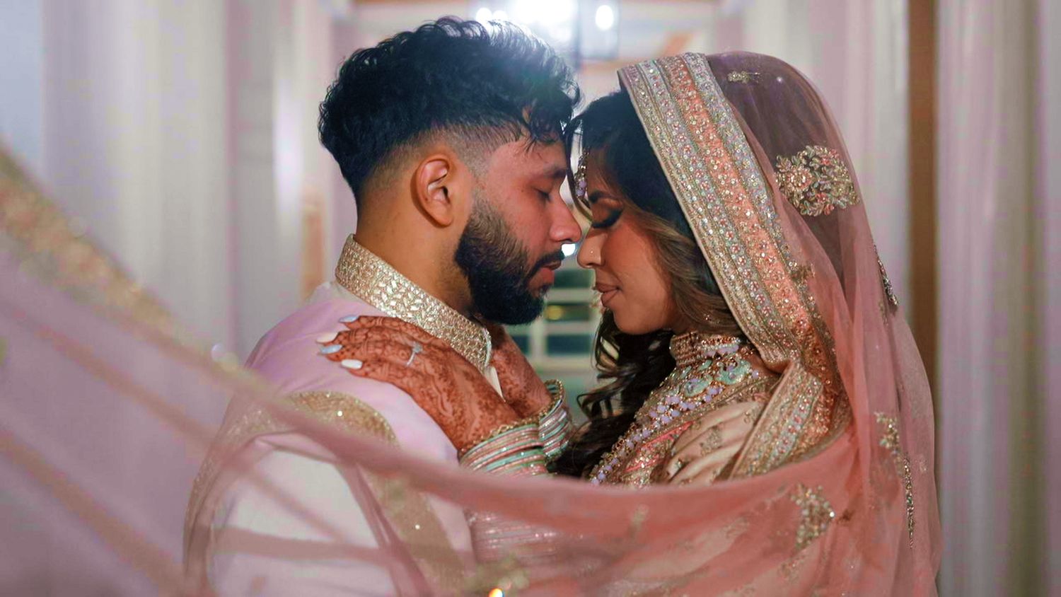 Indian bride and groom in pink traditional wedding attire share romantic forehead touch.