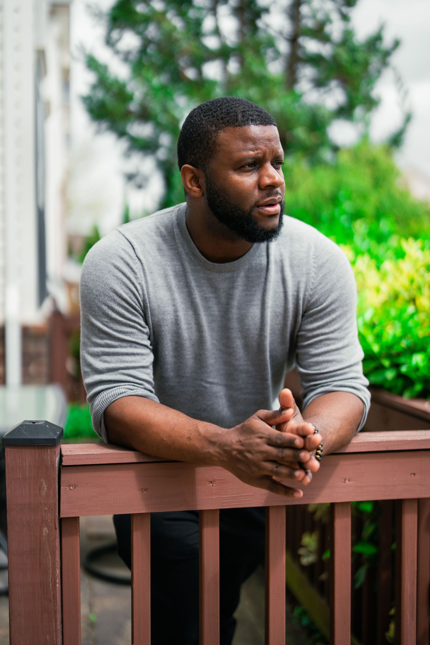 A muscular Black man in a gray sweater leans on a wooden deck railing, gazing thoughtfully to the side.