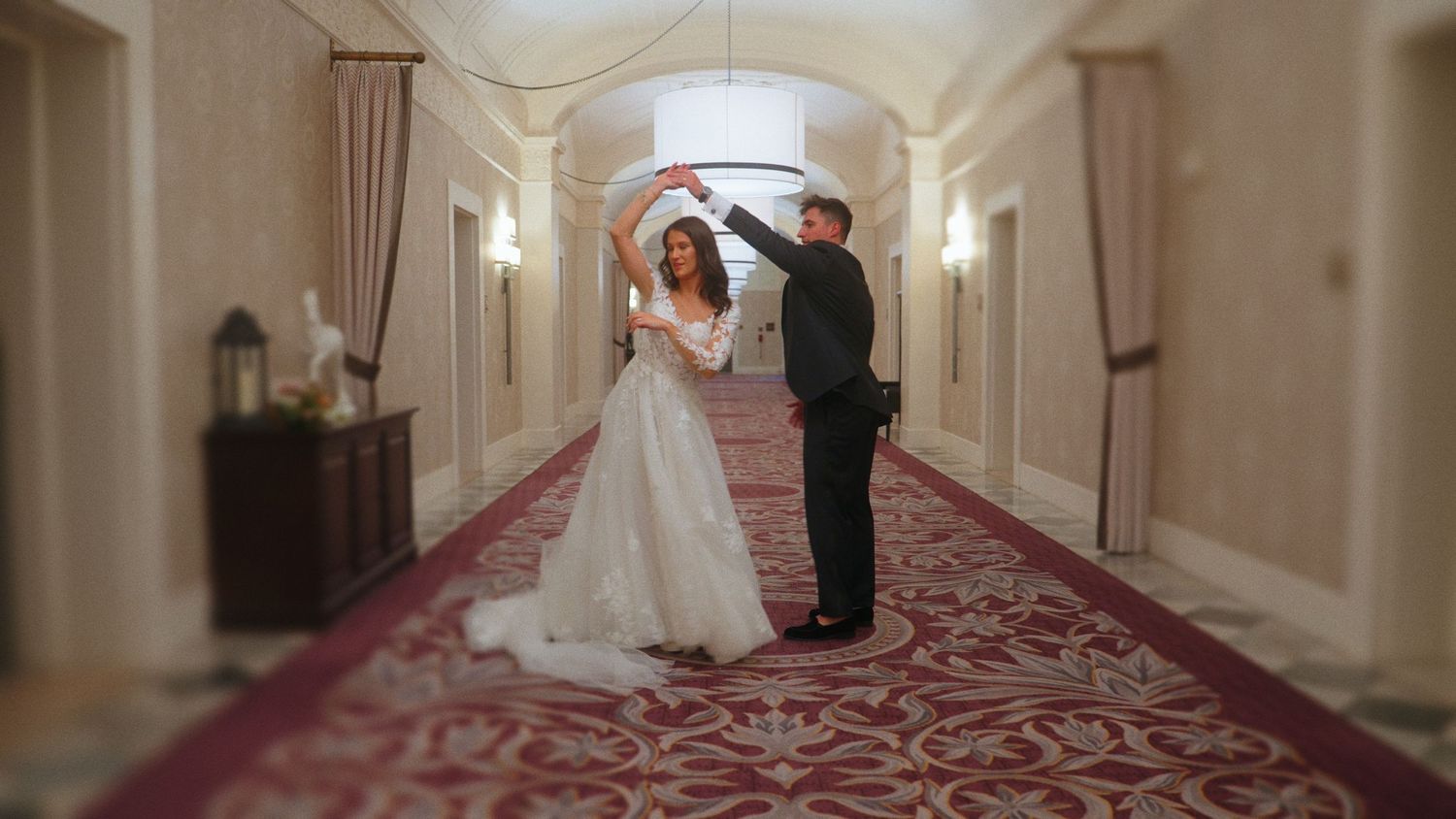 Bride and groom dancing together in an elegant hotel hallway with red carpet and ornate decor.