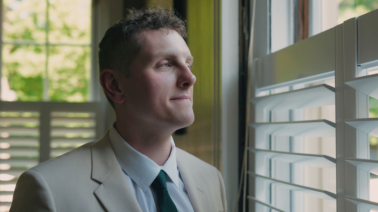 Handsome groom in beige suit gazes thoughtfully out window with natural light on his face.