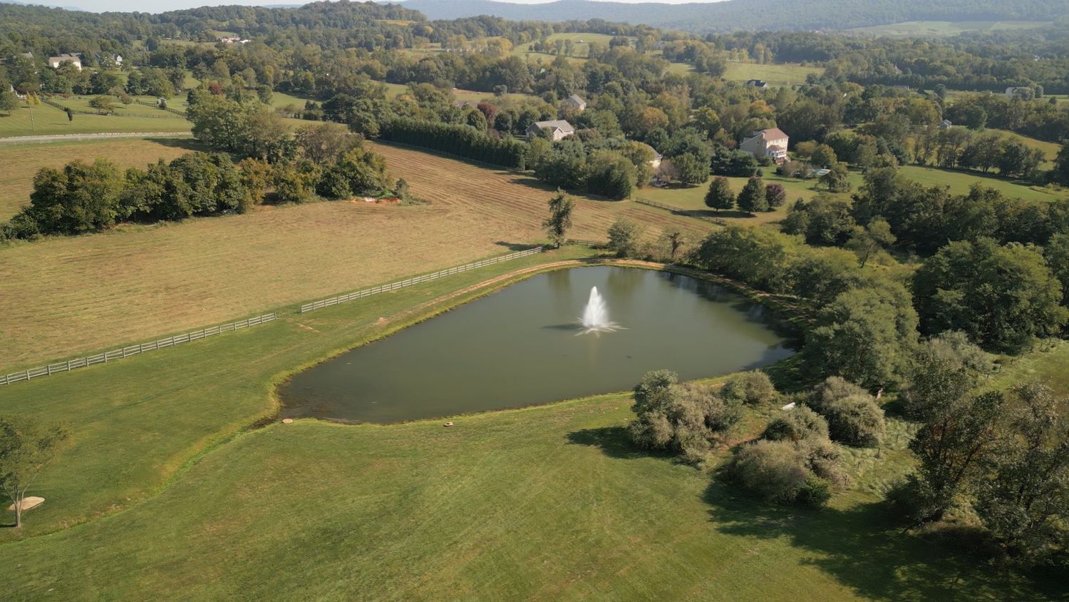 Aerial drone view of scenic countryside estate with pond and fountain surrounded by green fields.