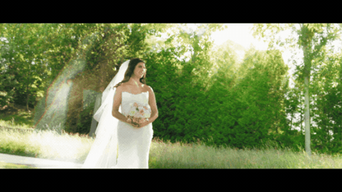 Bride in white gown and veil standing outdoors among lush green trees on her wedding day.