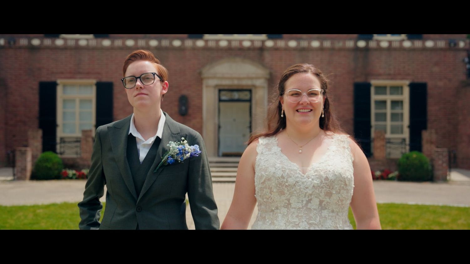Newlyweds hold hands and smile walking in front of a classic brick estate.
