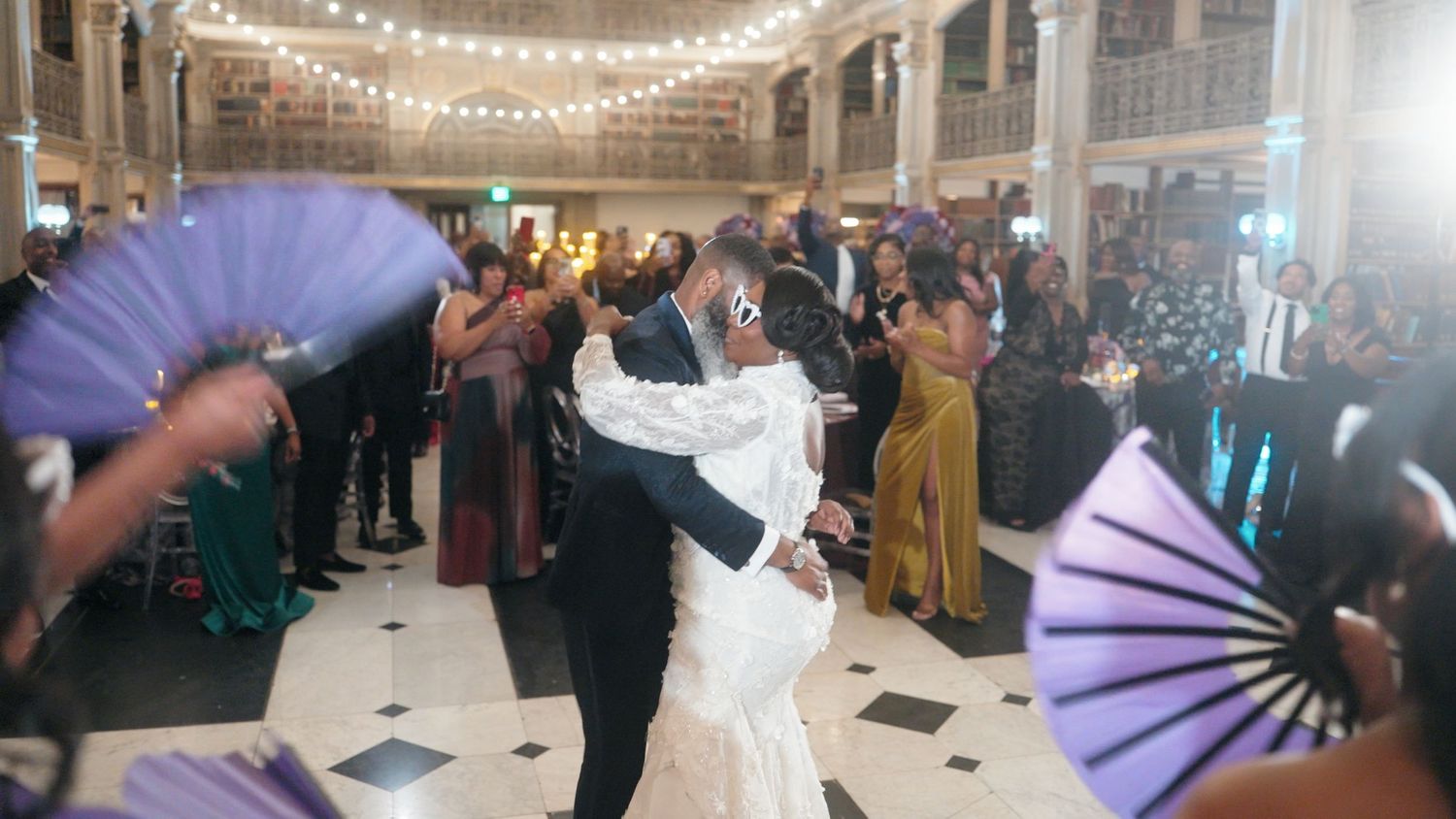 Bride and groom sharing first dance surrounded by guests waving purple fans at elegant wedding reception.