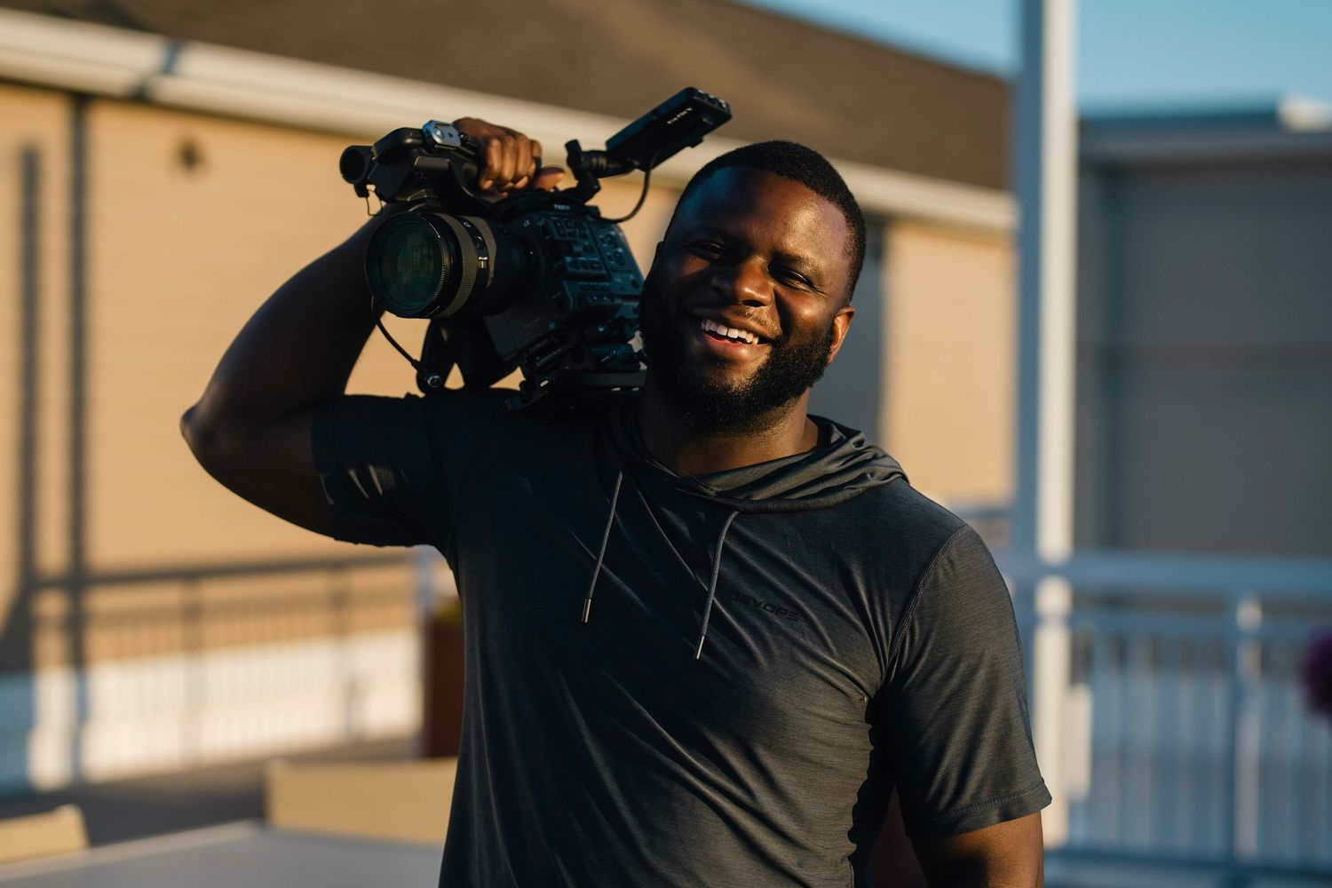 Smiling videographer holding a professional camera on his shoulder in golden hour sunlight.