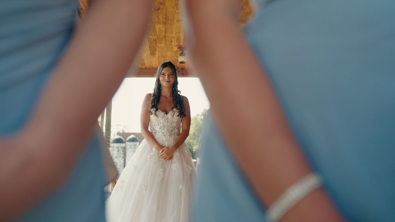 Bride in white floral wedding gown viewed through bridesmaids in blue dresses.