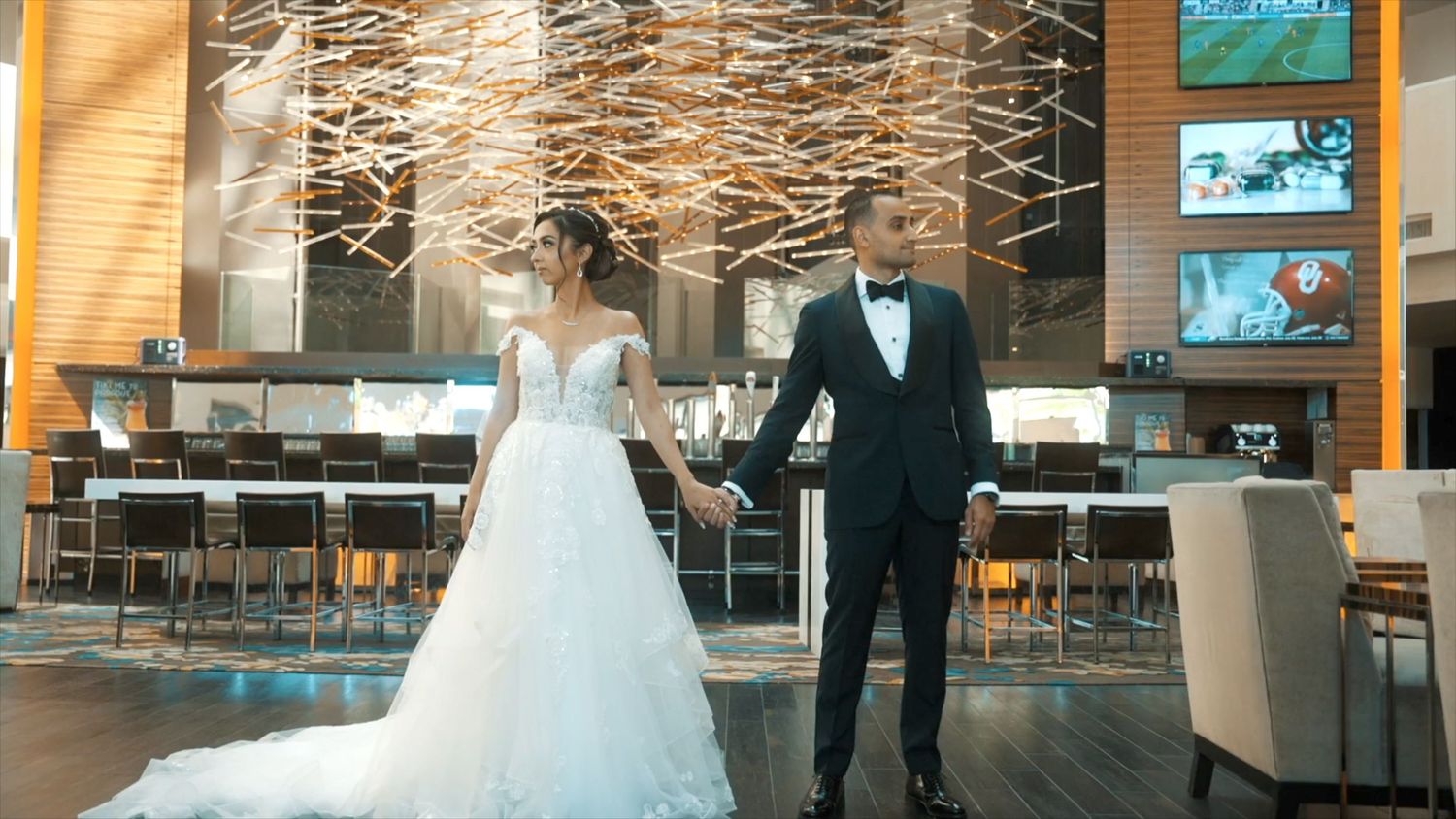 Bride in ball gown and groom in tuxedo hold hands in grand hotel lobby with sparklers.