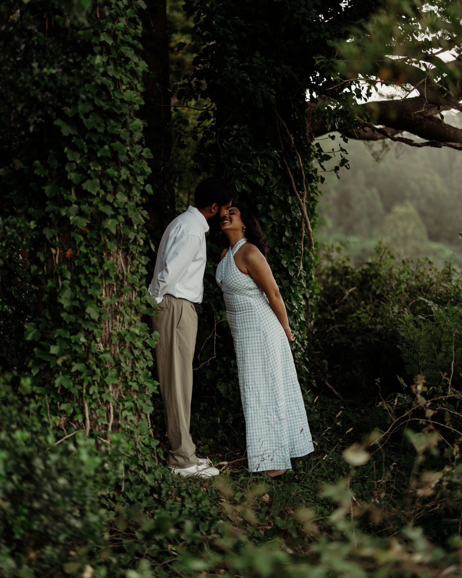 A couple shares a romantic moment among ivy-covered trees in a lush green forest setting.