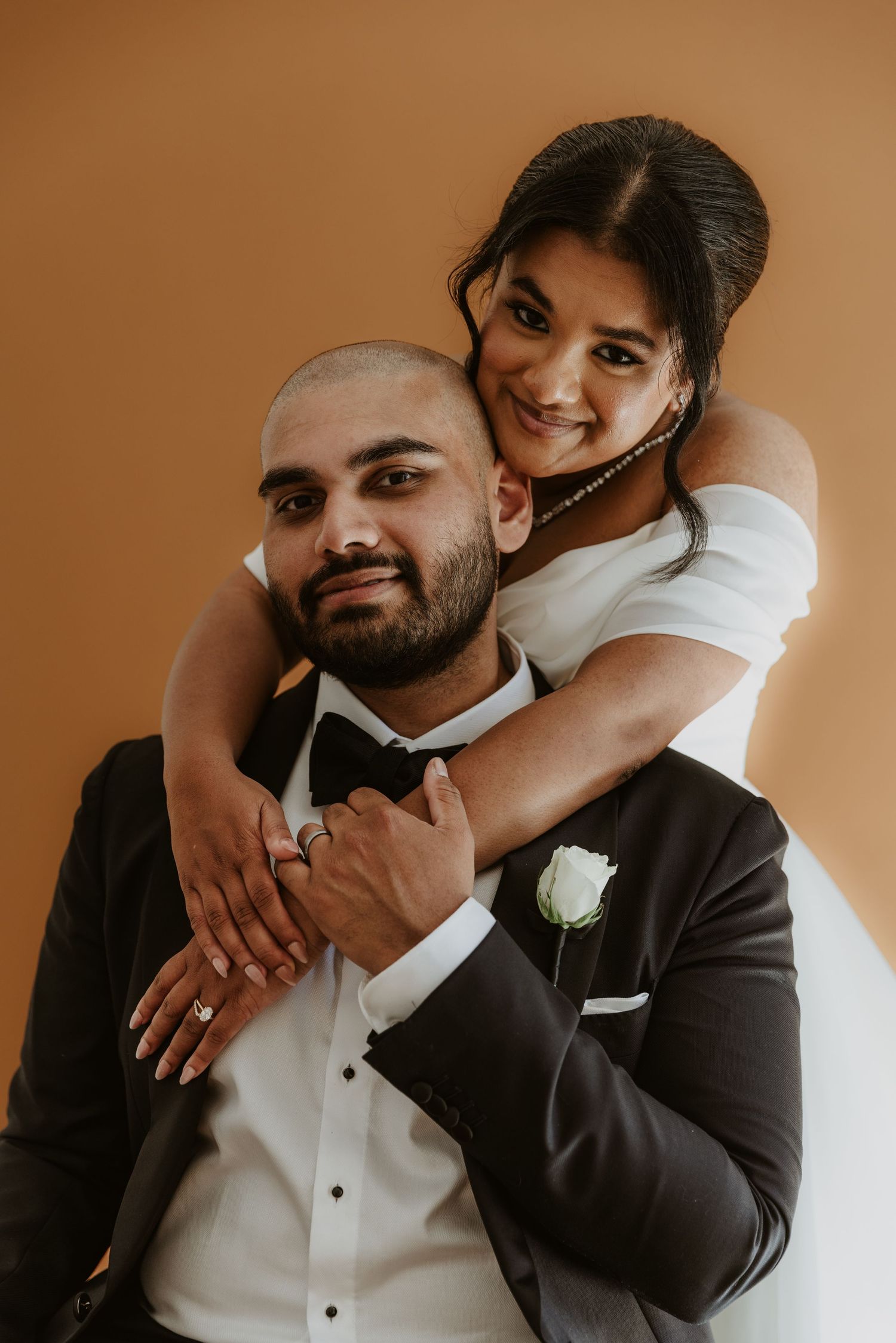 Romantic couple portrait in formal attire with white boutonniere against warm brown background.