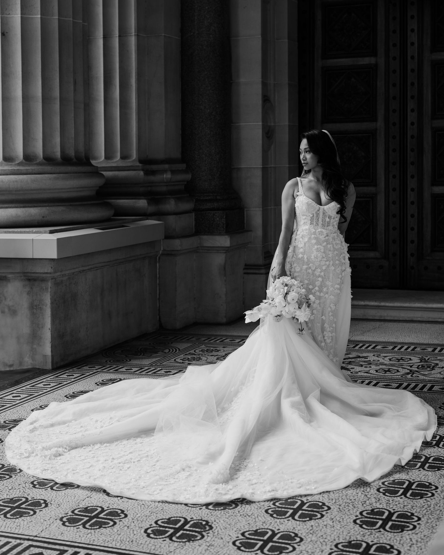 Black and white bridal portrait of a bride in a lace mermaid gown with train, holding bouquet beside ornate columns.