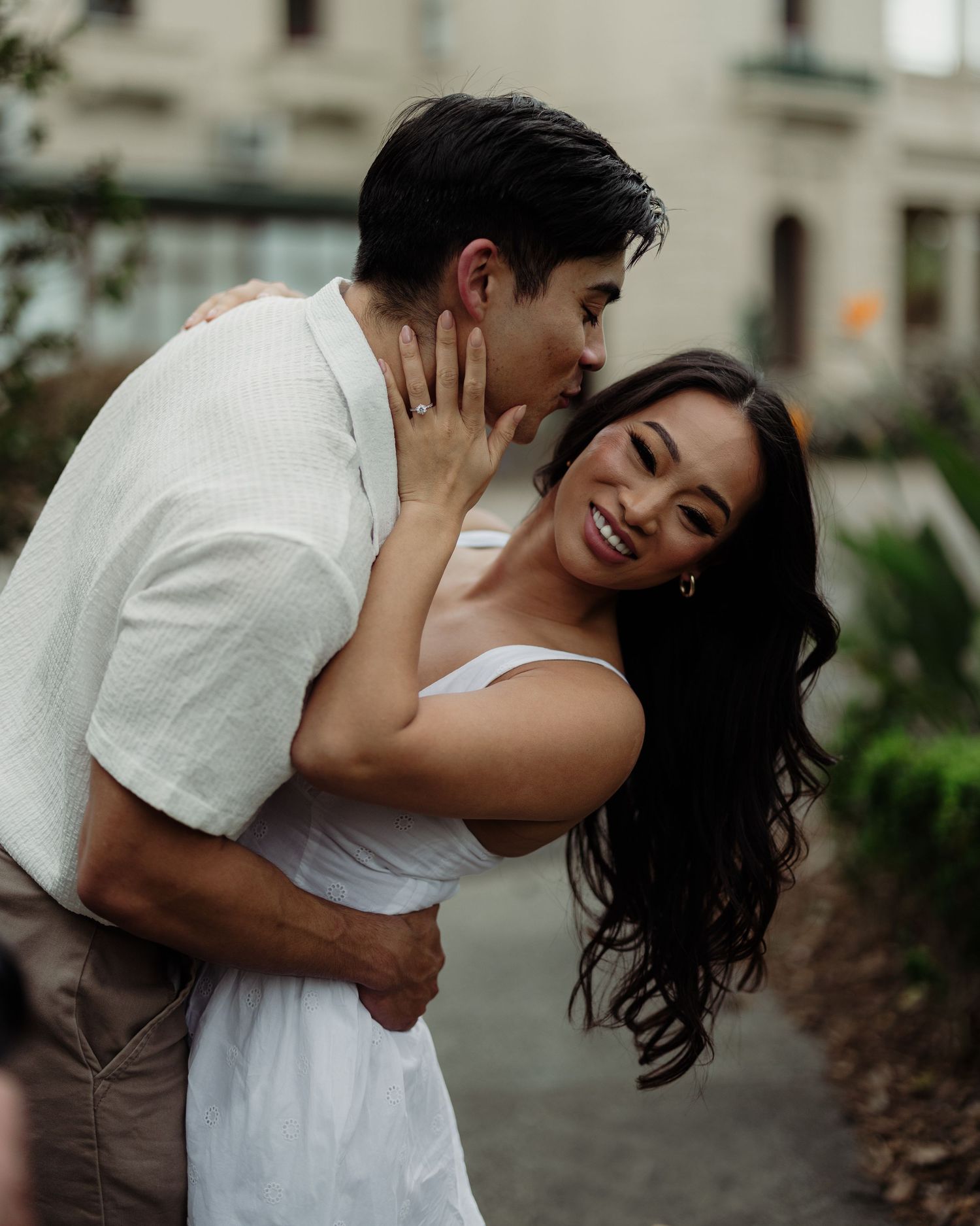 Romantic couple in white clothing share an intimate moment against an urban backdrop.