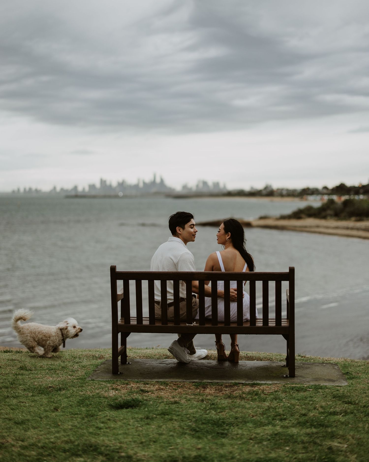 Couple sits together on wooden bench overlooking ocean with their dog nearby.