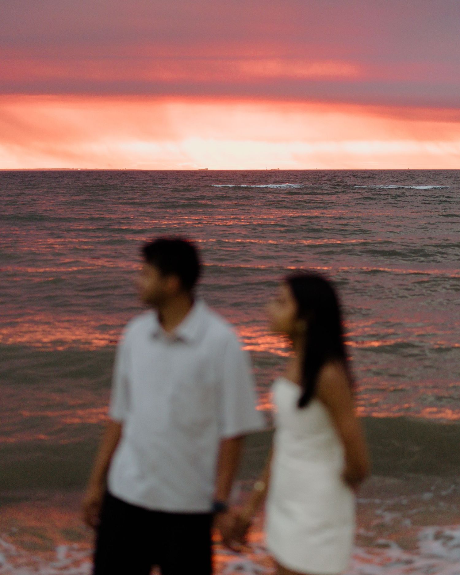 Couple walking along beach at sunset with dramatic pink and orange sky reflecting on ocean waves.
