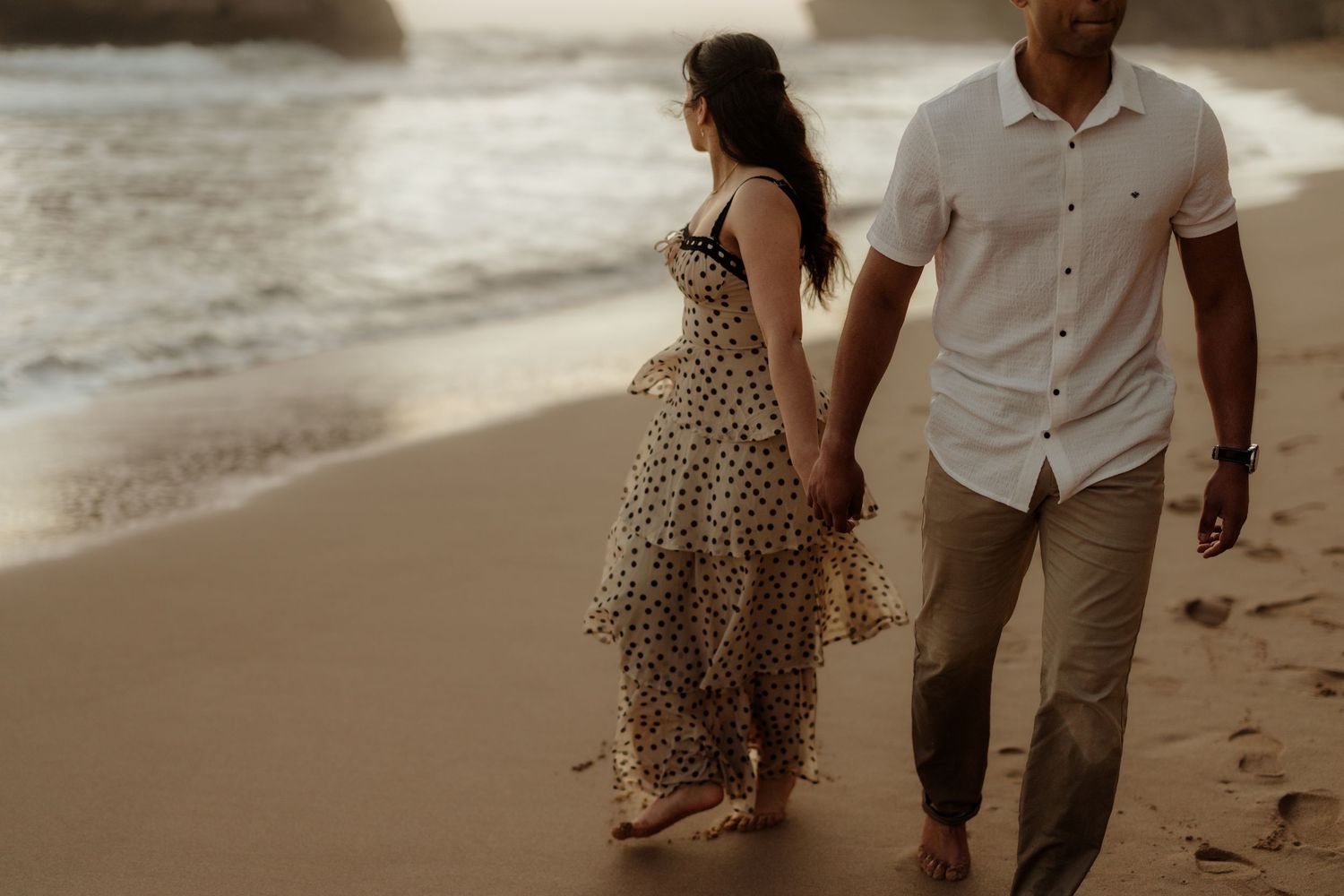 Couple holding hands walking barefoot on a sandy beach at sunset, woman in polka dot dress, man in white shirt.