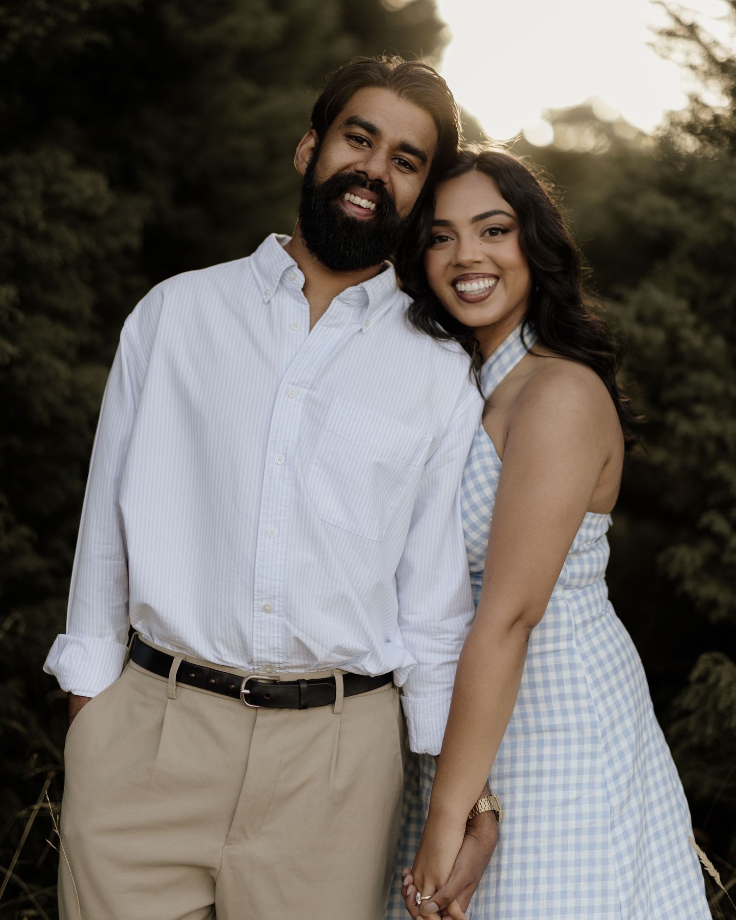 A smiling couple poses outdoors at golden hour, man in white shirt and khakis, woman in blue gingham dress.