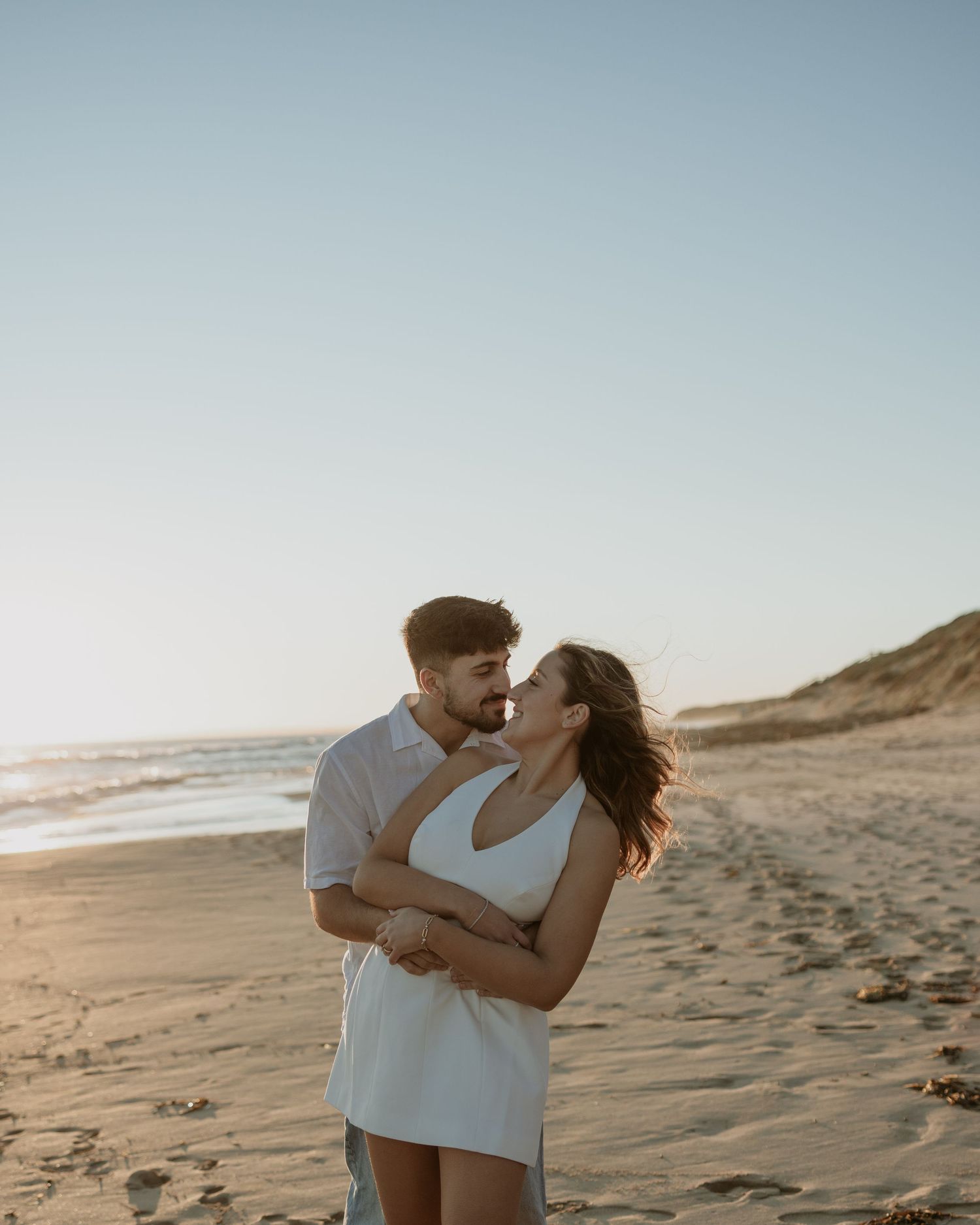 A romantic couple embraces on a sandy beach at sunset with golden light illuminating their silhouettes.