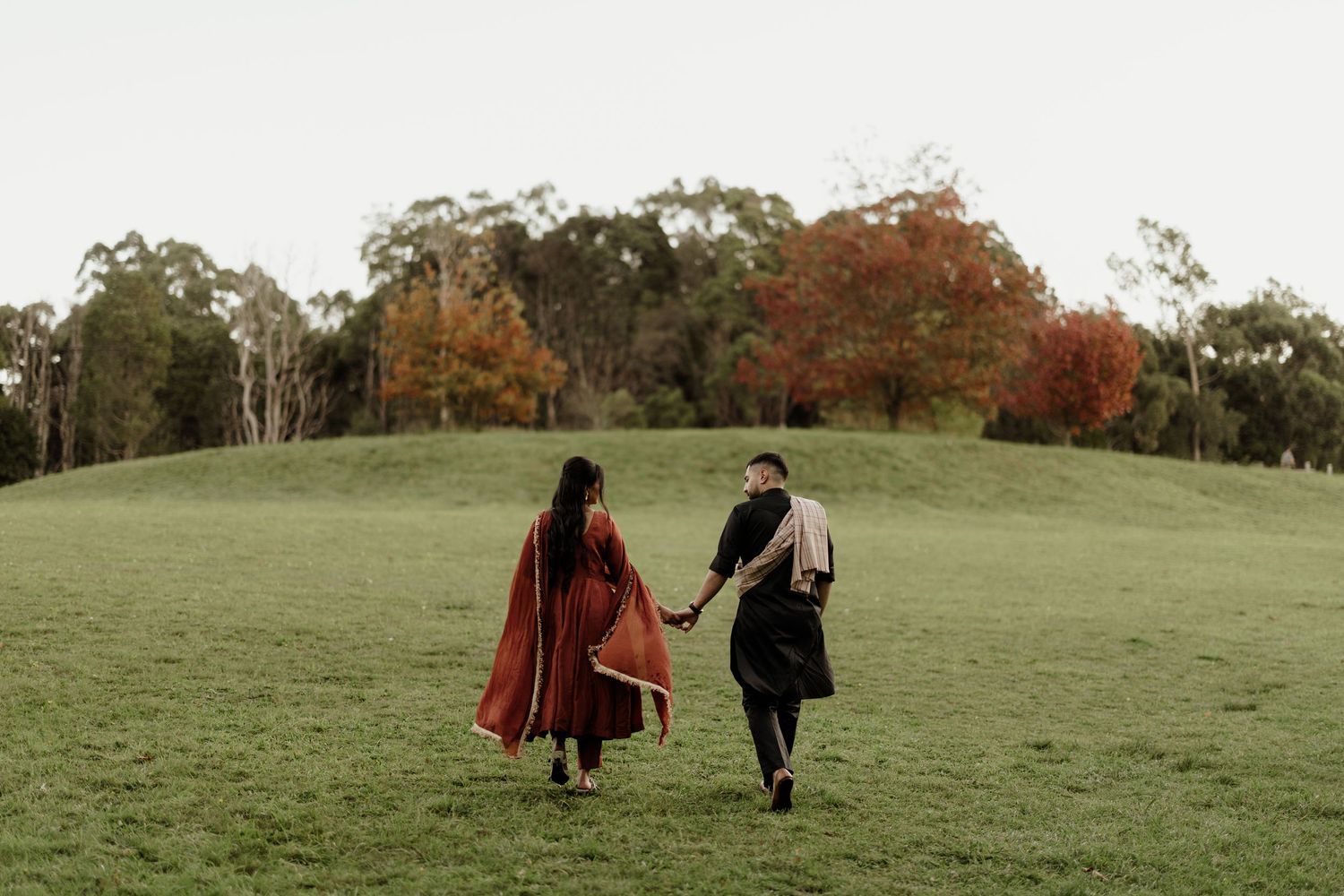 Couple holding hands walking across lush green field with colorful autumn trees in background.