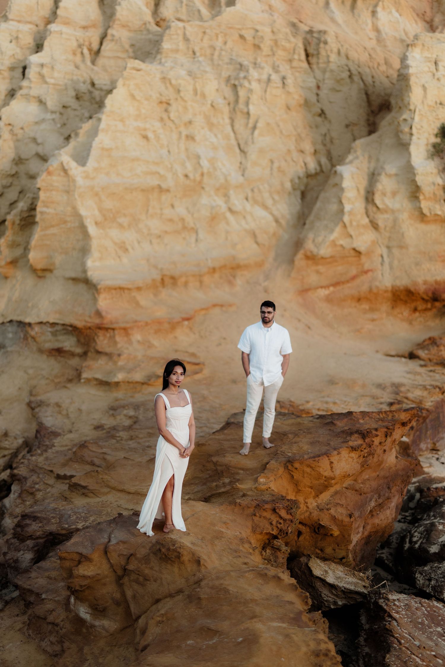 Couple in white outfits posing on rocky terrain with golden sandstone cliffs in the background.