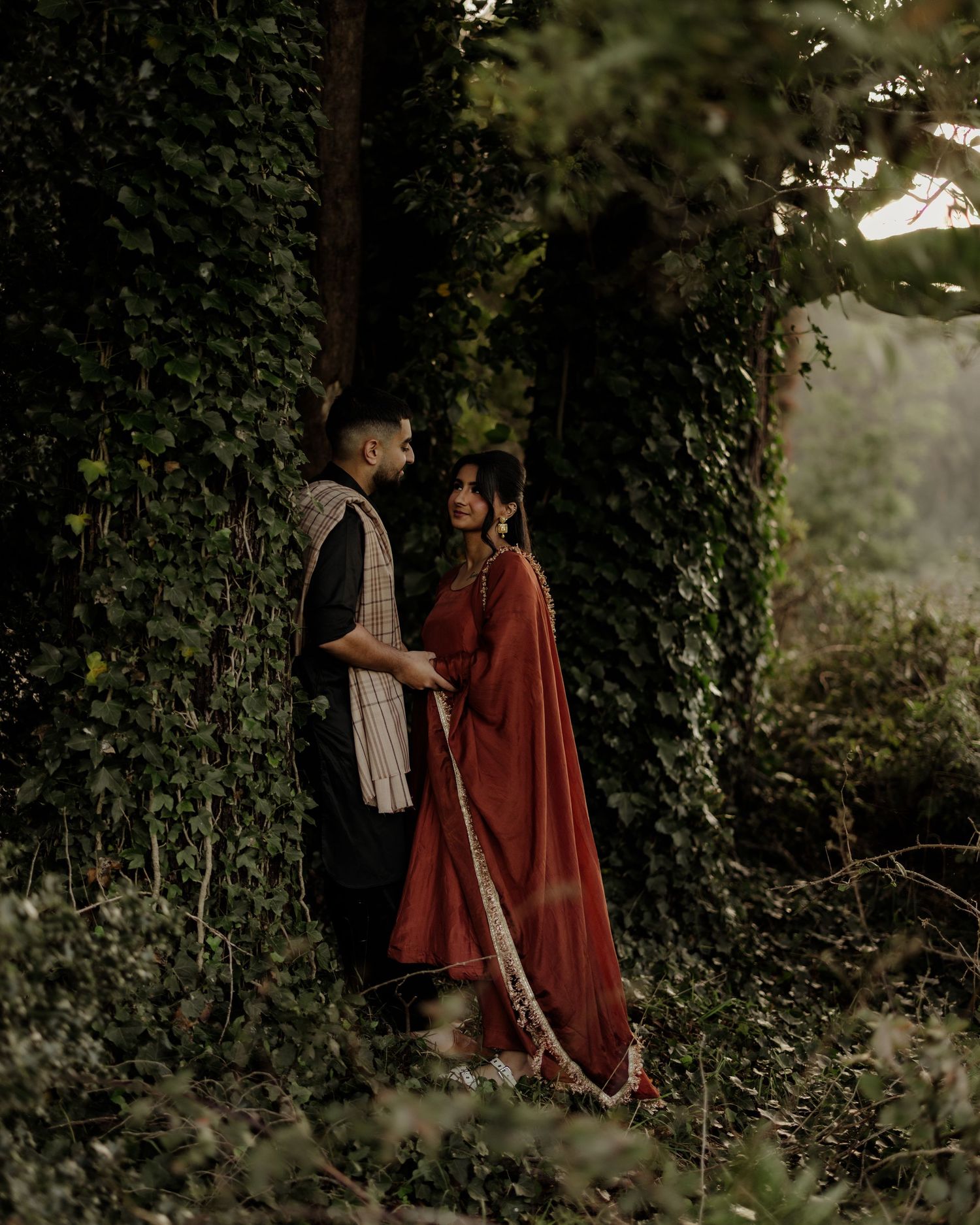 A couple in traditional South Asian attire stands romantically among lush ivy-covered walls in a moody outdoor setting.