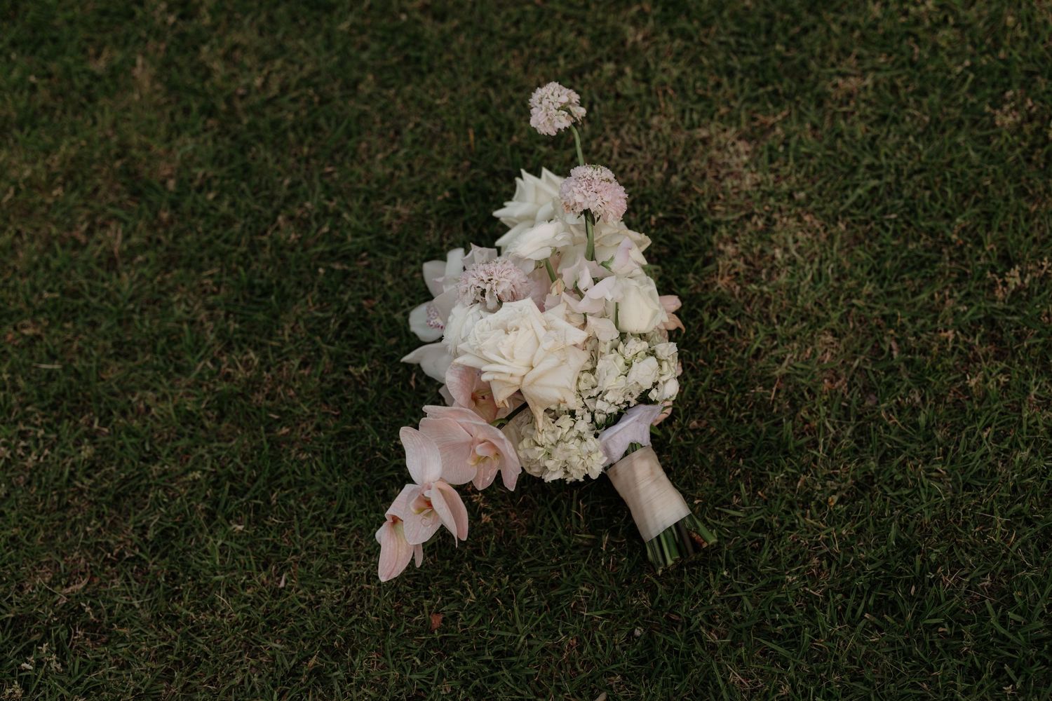 Delicate white and blush wedding bouquet with roses and hydrangeas lying on grass.