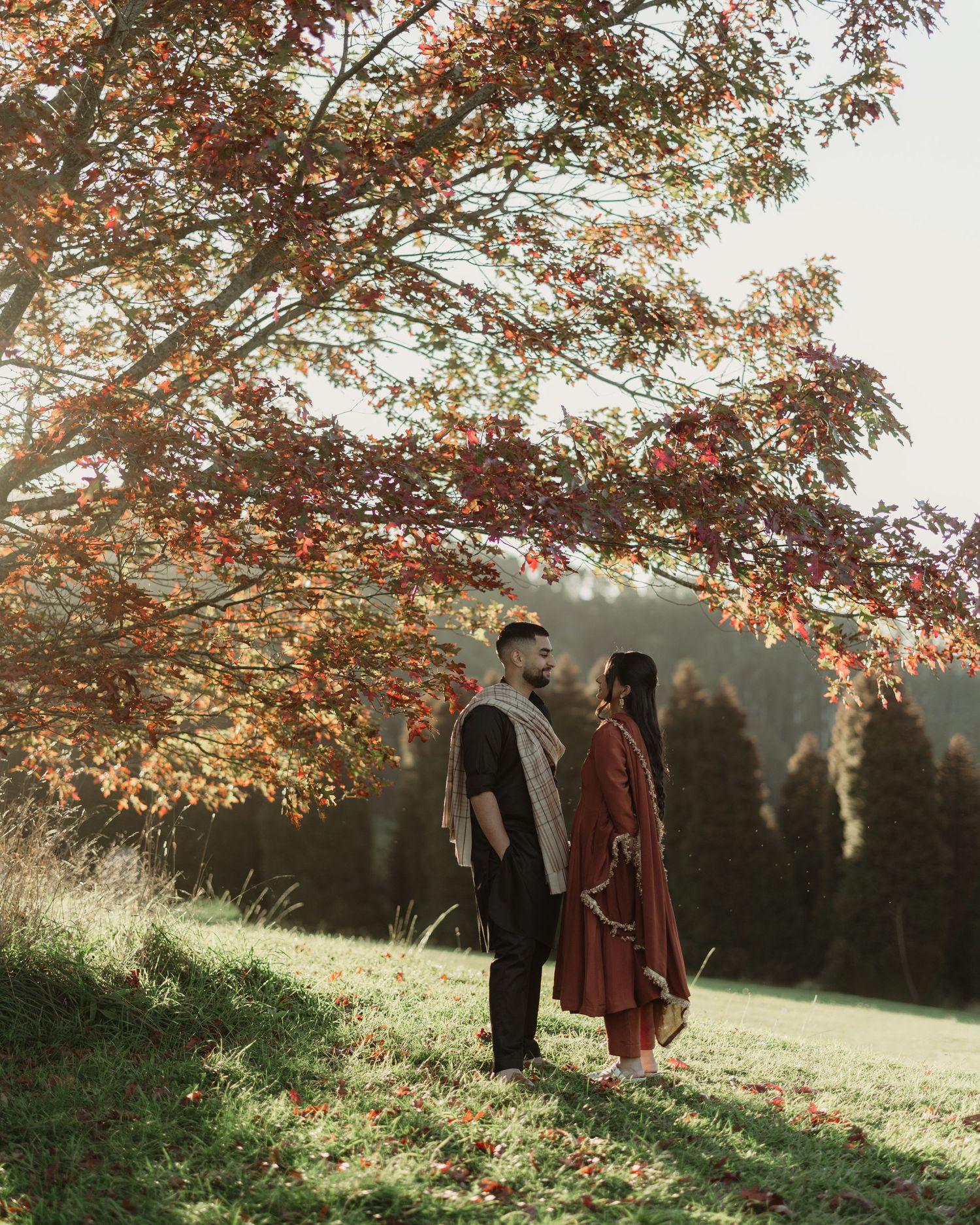 Couple standing face to face under a vibrant autumn tree on a green hillside with misty mountains in background.