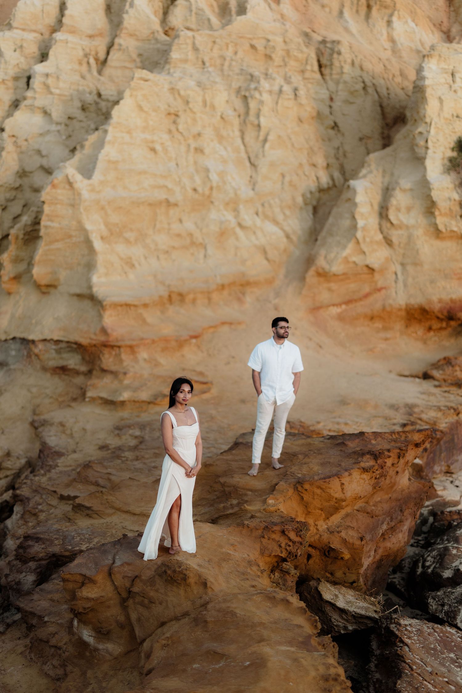 Couple in white outfits posing on rocky terrain with stunning sandstone cliffs in the background.