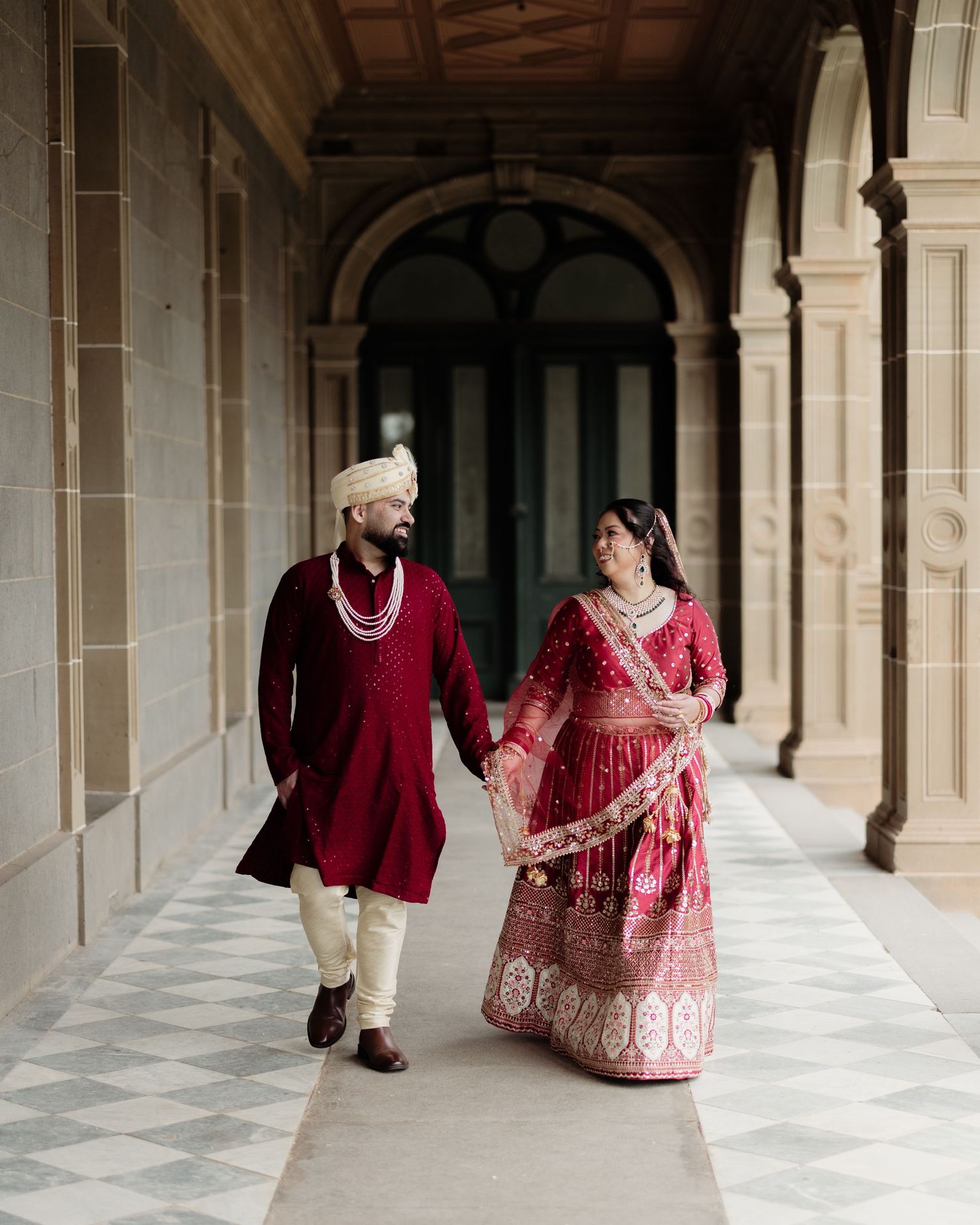 Indian couple in matching red traditional wedding attire walking hand in hand through elegant archway corridor.