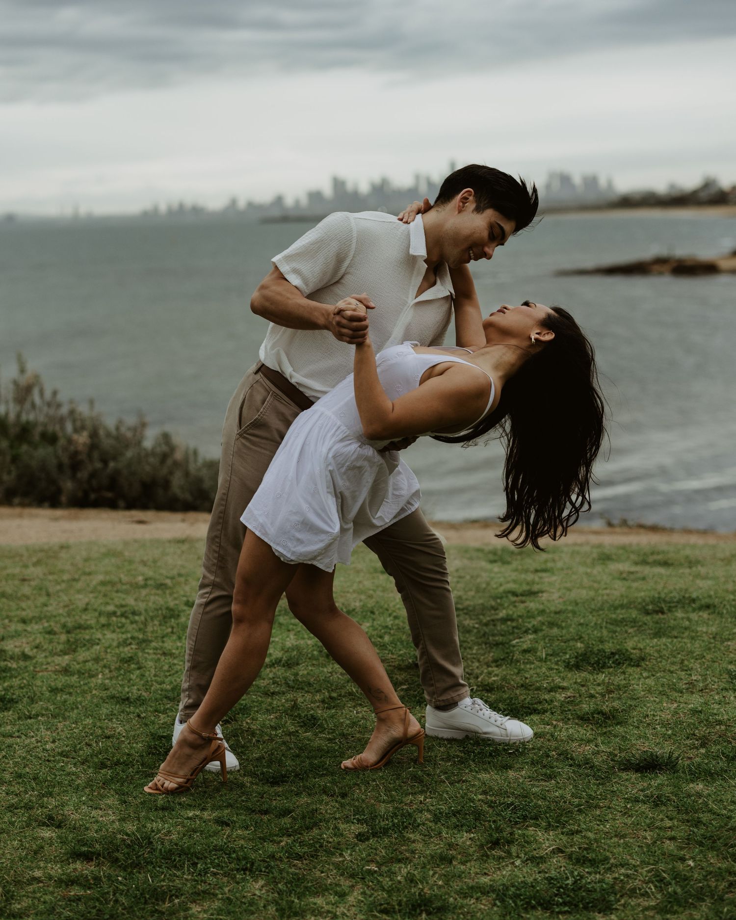 Playful couple performs a dramatic dance dip on grass overlooking the ocean.