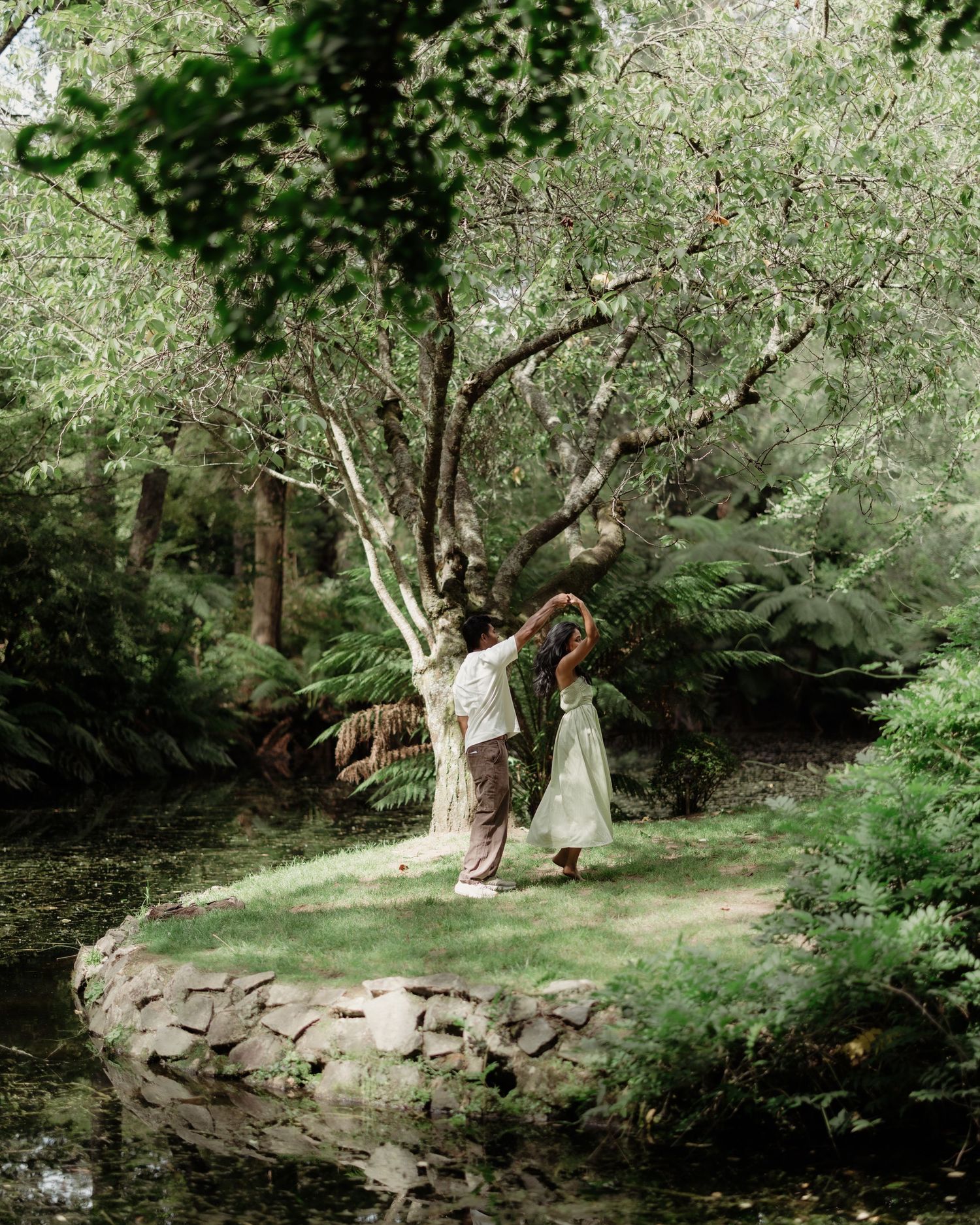 Couple dancing together in a lush green forest garden beside a tranquil pond surrounded by ferns and trees.