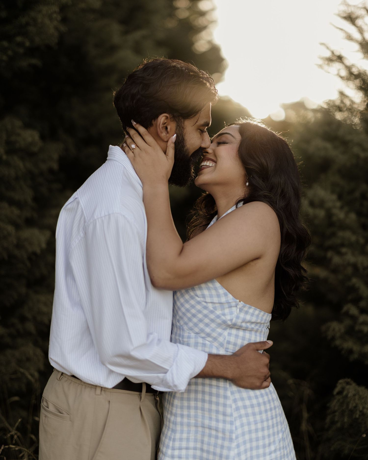 Couple sharing a romantic moment outdoors at golden hour, woman smiling in gingham dress, man in white shirt.