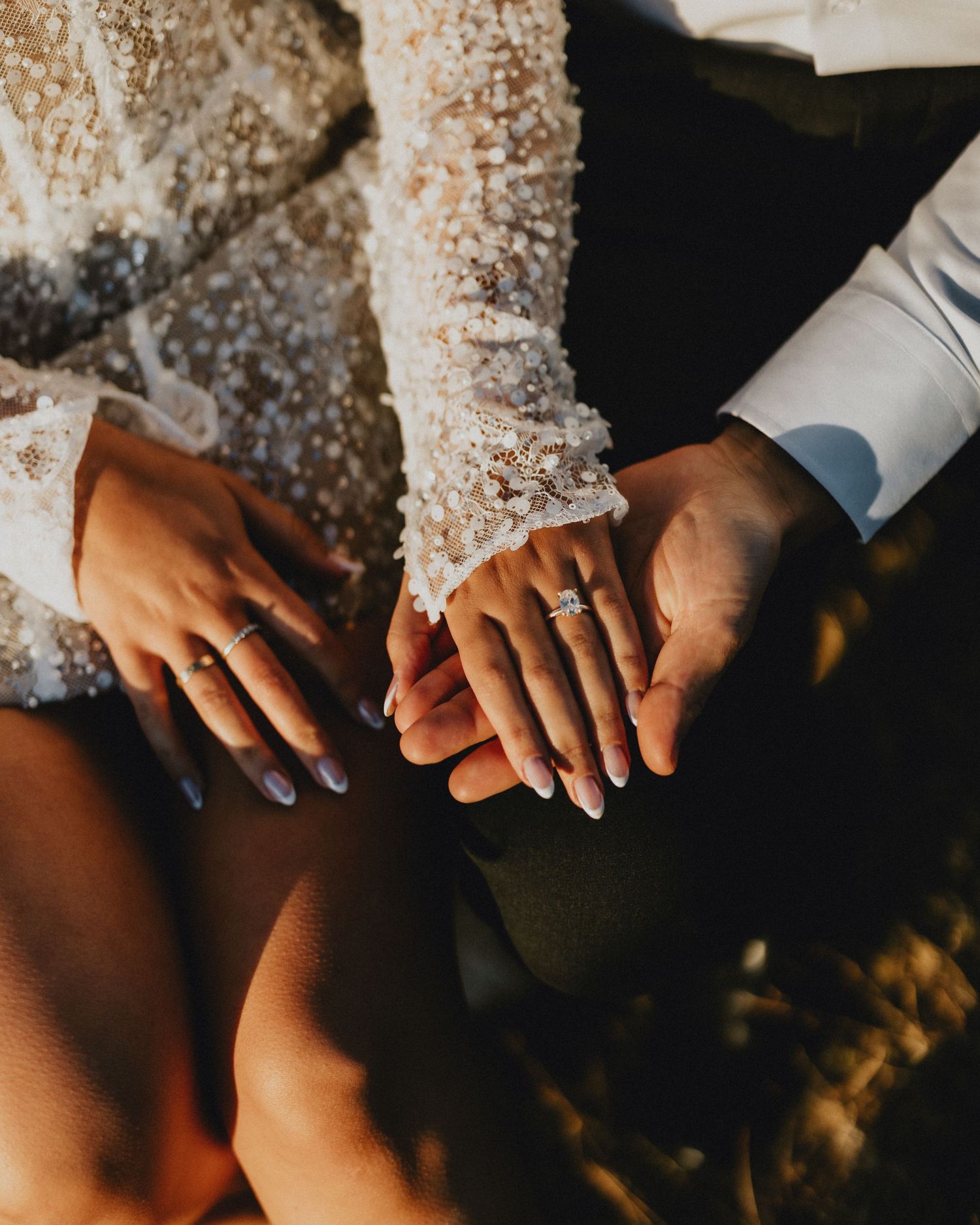 Hands with wedding rings touching intimately against white lace dress in warm sunlight.
