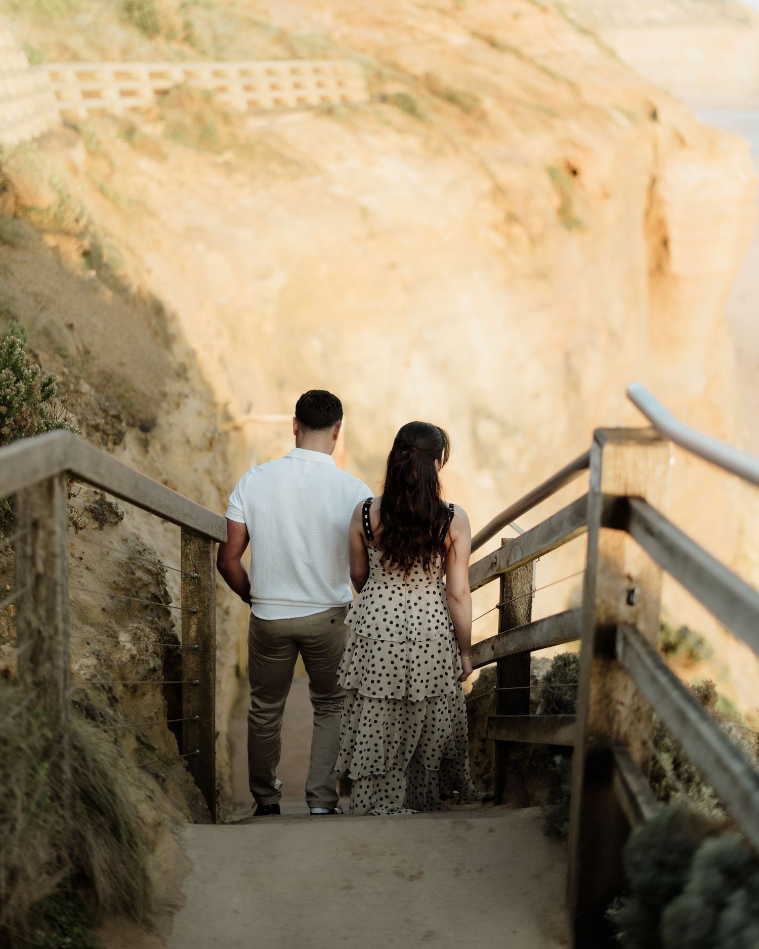 Couple holding hands on coastal cliffside stairs overlooking sandy bluffs, woman in polka dot dress.